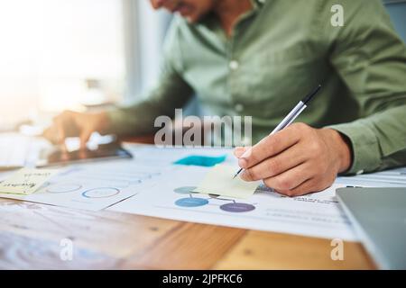 The business wont survive without him. an unrecognizable businessman making notes on a paper while browsing on a digital tablet at his desk in the Stock Photo