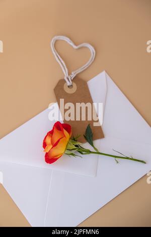 Beautiful red roses flowers in postal envelope on neutral beige background, beige paper note with rope in heart shape copy space for text, spring time, greeting card for holiday. Flower delivery. Delicate red yellow roses. Minimal trendy composition. Romantic pastel flowers. Neutral earth tones Stock Photo