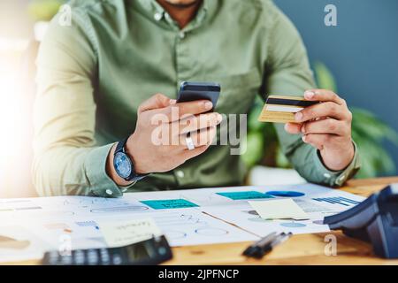 Time to make some payments. an unrecognizable businessman seated behind his desk while holding his credit card and cellphone inside of the office. Stock Photo