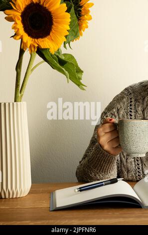 Woman with coffee cup in her hand. Notebook, pen and vase with sunflowers on the wooden table. Concept of inspirational workplace and home office. Stock Photo
