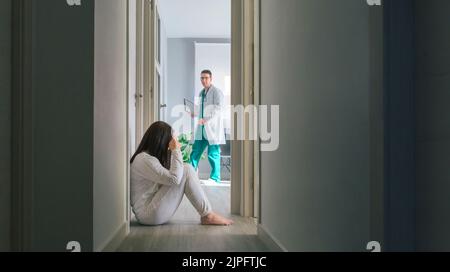 Doctor looking at female patient with mental disorder sitting on floor in hospital Stock Photo