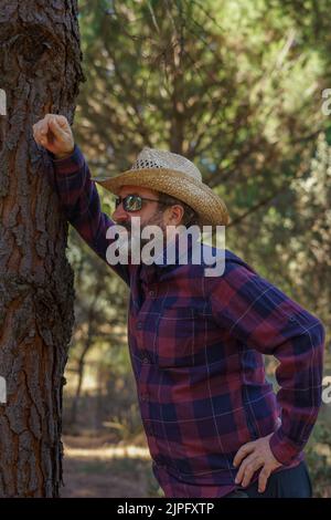 close-up of a man with a beard, hat and sunglasses leaning against a pine tree in the forest Stock Photo