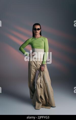 Full length of trendy nonbinary model in sunglasses holding jacket on abstract background Stock Photo