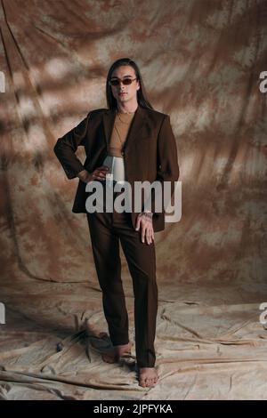 Full length of stylish queer person in suit and corset standing on abstract brown background Stock Photo