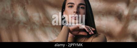 Portrait of young tattooed queer person looking at camera on abstract brown background, banner Stock Photo