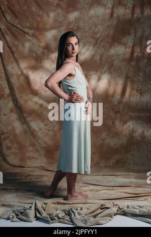 Full length of barefoot queer person in sundress looking at camera on abstract brown background Stock Photo