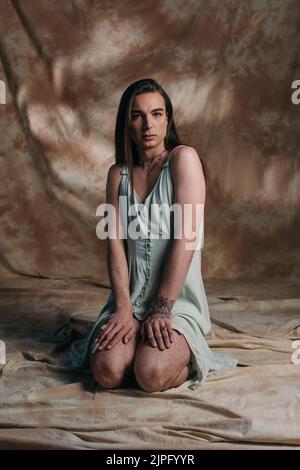 Young tattooed queer person looking at camera while sitting on abstract brown background Stock Photo