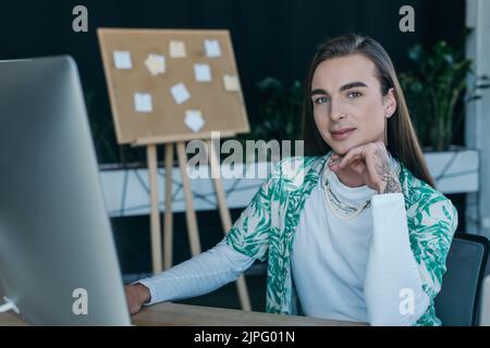 Tattooed queer designer looking at camera near computer in creative agency Stock Photo