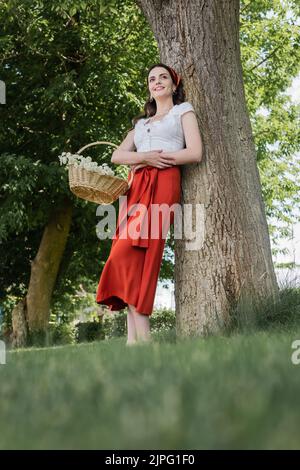 Low angle view of smiling woman in blouse and skirt holding basket with flowers in park Stock Photo