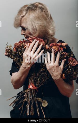 Sad elderly woman touching bouquet of withered rose flowers. Gray haired mature lady in depression. Concept of old aging. Studio shoot isolated on gray background. Copy space Stock Photo