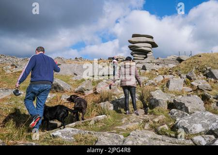 Dog walkers climbing up Stowes hill towards towering granite rock stack The Cheesewring left by glacial action on Bodmin Moor in Cornwall. Stock Photo
