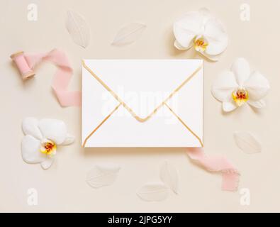 Paper envelope near white orchid flowers and light pink silk ribbons on light beige, top view, Wedding mockup. Romantic scene with horizontal blank ca Stock Photo