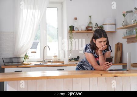 A young woman is in the kitchen in her house. The woman looks thoughtfully to the smart phone, determining what she needs to cook for dinner, there is a laptop next to her. High quality photo Stock Photo