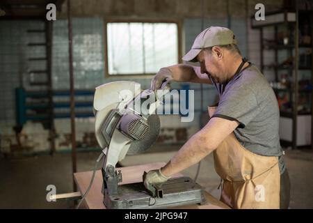 Worker saws metal. Guy in garage cuts steel with saw. Man uses electric tool. Guy works. Stock Photo