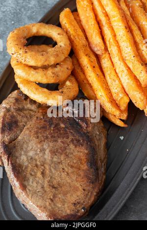 Rump steak dinner, with sweet potato fries and onion rings, on a black oval plate.  On a dark stone background Stock Photo