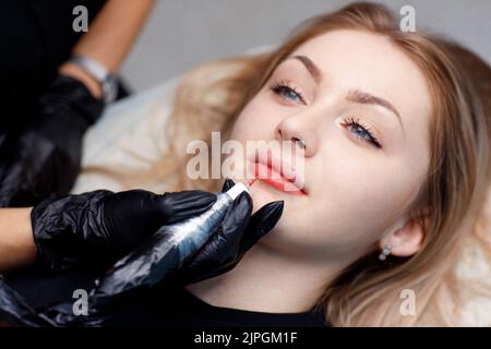 Cosmetologist applying red permanent make up tattoo on young woman lips. Stock Photo