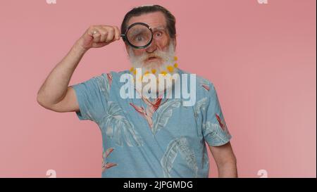 Investigator researcher scientist senior man with flowers in beard holding magnifying glass near face, looking into camera with big zoomed funny eye, searching, analysing. Elderly grandfather indoors Stock Photo