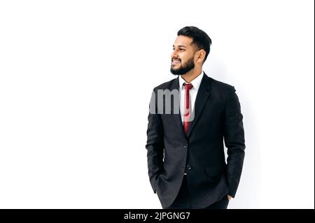 Handsome elegant influential arabian or indian man in formal suit, standing on isolated white background, looking to the side, thinking, planning, dreaming, smiling. Copy-space for your presentation Stock Photo