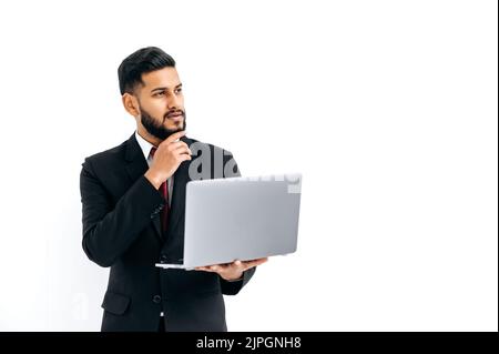 Puzzled thoughtful elegant influential arabian or indian man in formal suit, stand on isolated white background with laptop, pensively looking away, thinking, dreaming.Copy-space for your presentation Stock Photo