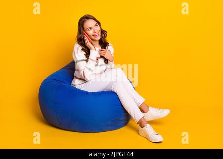 Full size photo of young pretty cute lady sitting pouf talking telegram call look empty space isolated on yellow color background Stock Photo