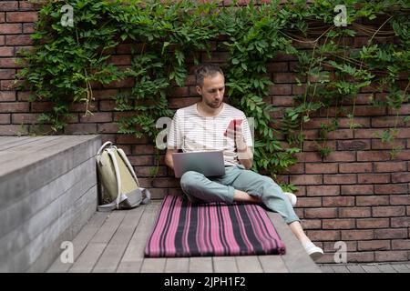 European student guy sitting with laptop outdoors using smartphone, being distracted by mobile phone Stock Photo