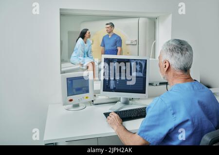 doctor radiographer waiting in control room behind protective glass while medical assistant prepares female patient for CT scan. Computed Tomography s Stock Photo