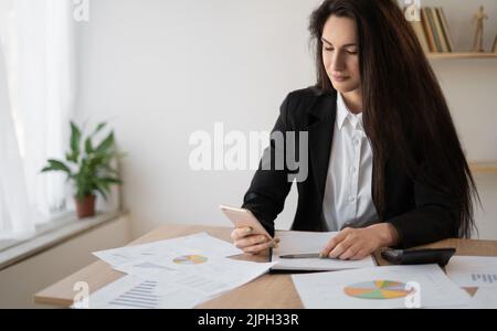 Beautiful hispanic woman working in office with mobile phone planning, accounting and data analysis. Business woman checking planner on smart phone Stock Photo