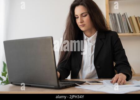 Portrait of a beautiful woman data analyst sitting at a desk working on a laptop. Stylish woman in an office setting. Searching for solution Stock Photo