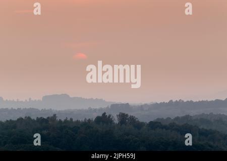 Atmospheric landscape scene of lush forest layers in mist during autumn sunset in countryside, forest obscured in fog and big orange sun dimmed behind Stock Photo