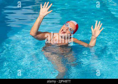 Middle aged white woman is having fun in the water of a swimming pool making a funny expression and wearing goggles and swimming cap Stock Photo