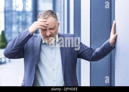 Senior gray haired man businessman upset and desperate outside office building, holding head with hands, mature boss bankrupt Stock Photo