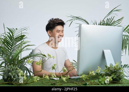 Happy excited Vietnamese young man working on computer with various plants around his workplace Stock Photo