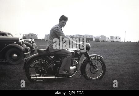 1950s, historical, a young gentleman sitting on a British made BSA motorcycle in a field at a caravan site, England, UK. Cars of the era parked up. Founded in 1861 as the Birmingham Small Arms Company Ltd, for the production of firearms, a motorcycle division of BSA was established in 1903, and the first motorcycle released in 1910. In the 1950s, BSA Motorcycles was the largest motorcycle maker in the world. Stock Photo