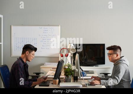 Two young Asian designers wearing eyeglasses using computers while wrapped up in work at spacious open plan office, profile view Stock Photo