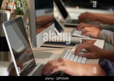 Working process at open plan office: group of unrecognizable programmers sitting at desks and finishing ambitious project with help of computers, close-up shot Stock Photo