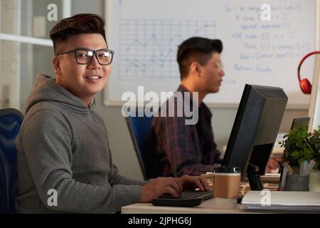 Smiling Asian student wearing eyeglasses looking at camera while sitting in front of modern computer during ICT class, portrait shot Stock Photo