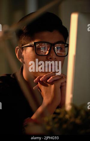 Pensive Asian manager wearing eyeglasses sitting in front of computer and analyzing statistic data while working late in dim office, portrait shot Stock Photo
