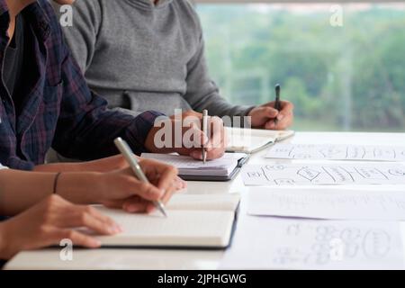 Close-up shot of young managers taking necessary notes while sitting at office desk and participating in working meeting Stock Photo