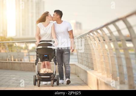 Attractive Caucasian woman kissing her Asian husband while pushing baby carriage and walking along waterfront, picturesque cityscape on background Stock Photo
