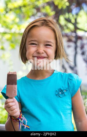 Unknown girl, wearing a green t-shirt, holding a milk chocolate ice cream, on the street, in summer, looking at the camera. Ice cream, popsicle, eatin Stock Photo