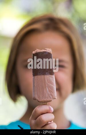 Unknown girl, wearing green shirt. out of focus, holding a milk chocolate ice cream, focused, on the street, in summer. Ice cream, popsicle, eating, s Stock Photo