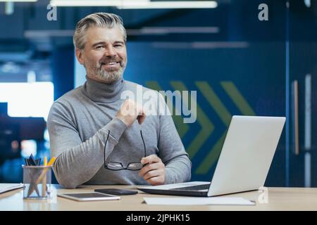 Portrait of senior gray-haired businessman, experienced mature man working with laptop inside office building, investor smiling and looking at camera Stock Photo