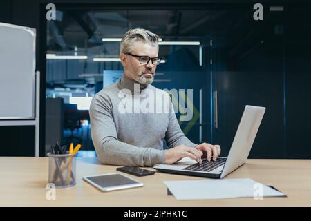 Experienced and mature successful businessman focused and serious man working inside office building at laptop, investor in glasses and casual clothes Stock Photo