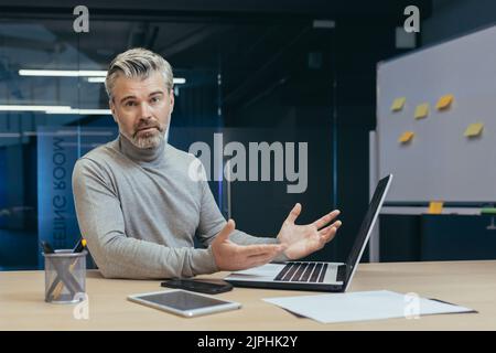 Portrait of upset gray-haired mature businessman, senior man looking at camera upset, businessman working in middle of office building using laptop Stock Photo