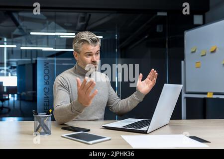 Frustrated and upset gray-haired experienced businessman, mature man spreading hands, negative work result, senior businessman at work using laptop Stock Photo