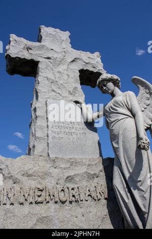 A stone angel stands with her hand on the cross and her wings partially spread with a look of sadness and loss against the dark blue sky Stock Photo