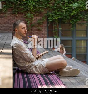 Thoughtful young man draws sketches sitting on stylized staircase step in park with people Stock Photo
