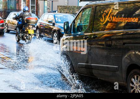 A van drives through a large roadside puddle caused by a mains water leak, showering the pavement with spray, London, UK Stock Photo