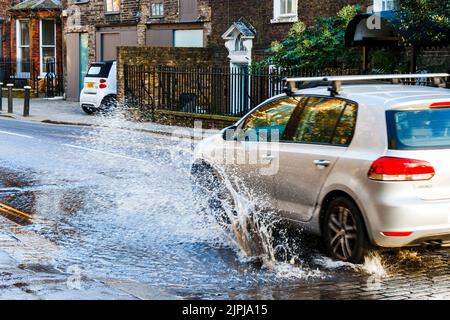 A car drives through a large roadside puddle caused by a mains water leak, showering the pavement with spray, London, UK Stock Photo