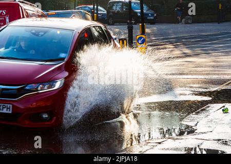 A car drives through a large roadside puddle caused by a mains water leak, showering the pavement with spray, London, UK Stock Photo